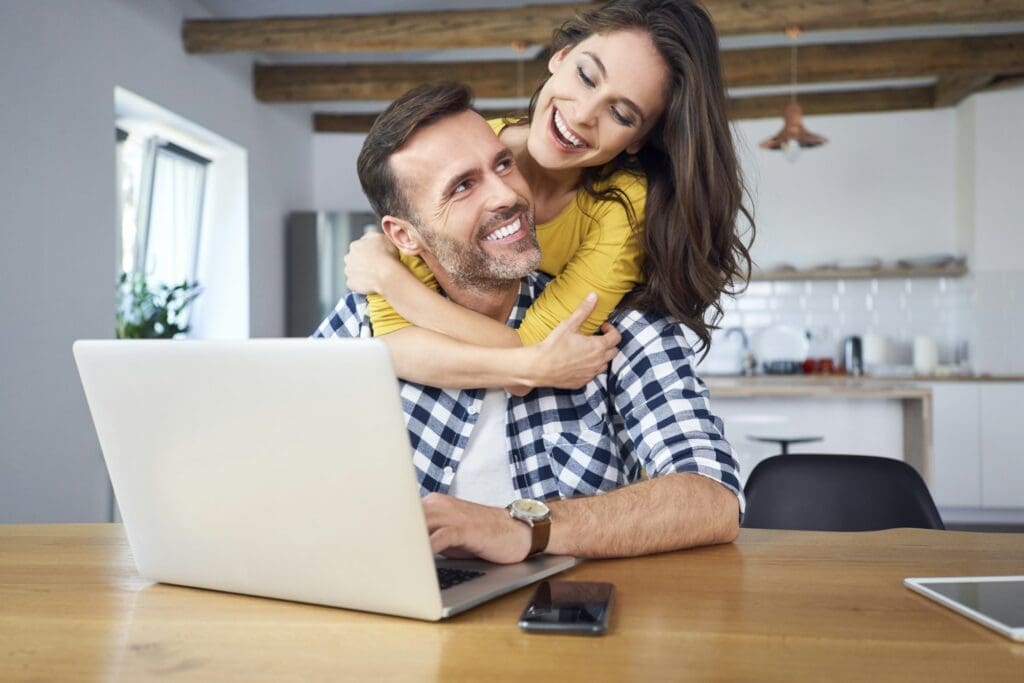 Happy couple sitting at dining table, embracing, using laptop